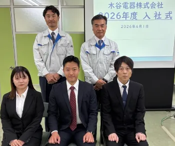 木谷電器の2026年度入社式。木谷社長、常務、そして3名の新入社員による記念写真。スクリーンには式典のスライドが投影され、社内の結束を示す一枚。