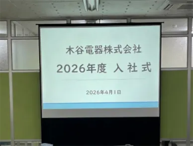 木谷電器株式会社の2026年度入社式スライド。「2026年度 入社式」「2026年4月1日」のテキストが投影された、オフィス内のプロジェクタースクリーン。